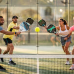 Qu’est-ce que le padel et comment se pratique ce sport de raquette ? Deux hommes et deux femmes jouent un échange intense de padel sur une piste extérieure vitrée, photographiés au niveau du filet sous une lumière naturelle.